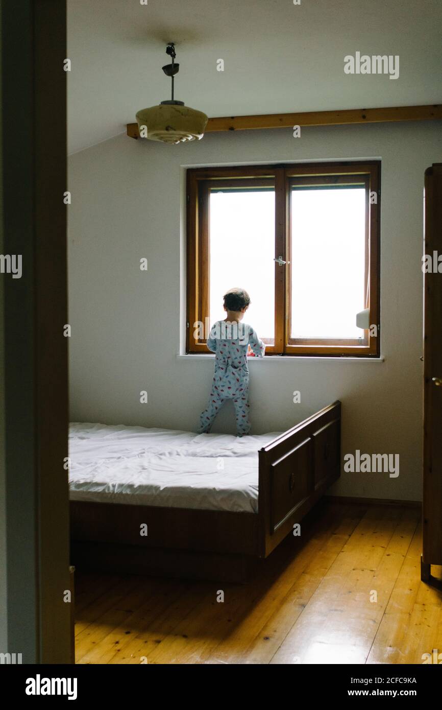 Rückansicht von kleinen Kind in bunten Schlafanzug Blick aus Fenster stehen auf dem Bett im Schlafzimmer Stockfoto