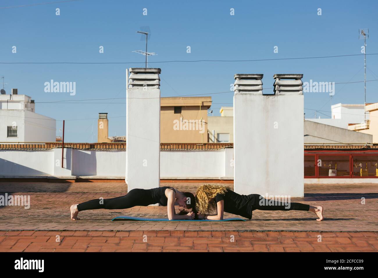 Seitenansicht der fokussierten Passform Frauen in schwarz activewear Durchführung Dolphin Plank oder Unterarm Plank Pose während Partner Yoga Praxis Auf der Dachterrasse bei sonnigem Tag in der Stadt Stockfoto