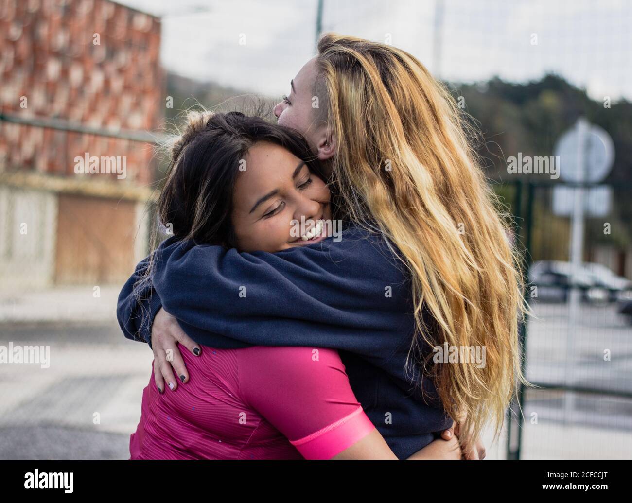 Seitenansicht von glücklichen, vielfältigen Sportlerinnen mit geschlossenen Augen stehend Einander umarmen und auf der Straße lachen Stockfoto