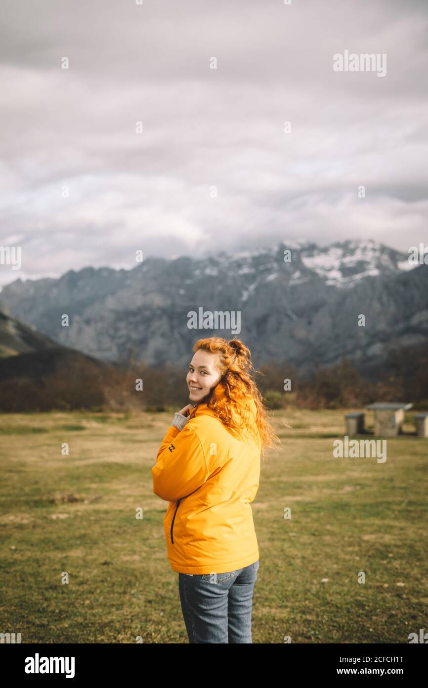 Rückansicht einer lächelnden Frau in einer warmen gelben Jacke, die die Kamera über der Schulter auf einer grünen Wiese, umgeben von verschneiten Bergen, anschaut Stockfoto
