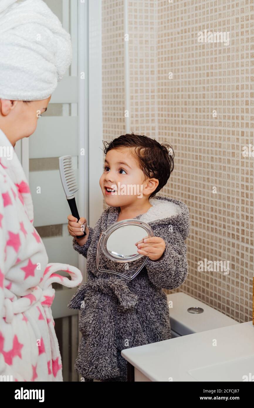 Liebenswert Kind im Bademantel stehen in hellen Badezimmer zusammen mit Mutter und Kämmen nasses Haar nach der Dusche beim Blick in Spiegel Stockfoto