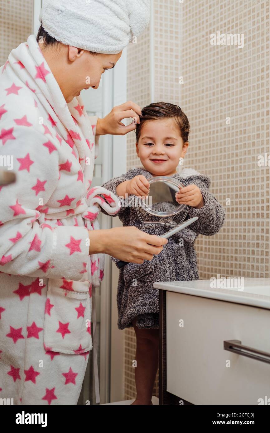 Seitenansicht der Mutter im Bademantel und Handtuch Turban Kämmen Nasse Haare von kleinen Kindern, während sie im modernen Badezimmer stehen Stockfoto