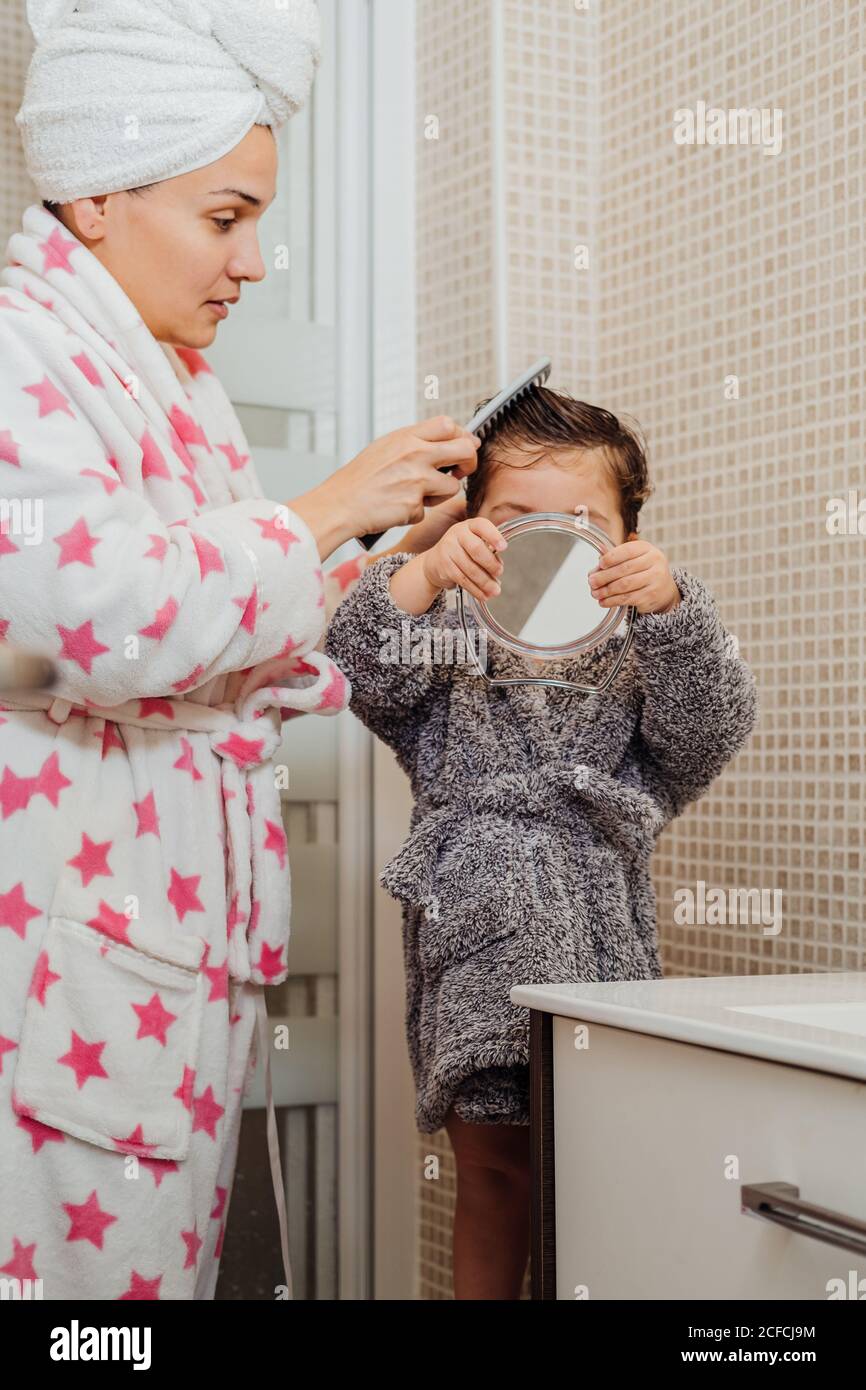 Seitenansicht der Mutter im Bademantel und Handtuch Turban Kämmen Nasse Haare von kleinen Kindern, während sie im modernen Badezimmer stehen Stockfoto