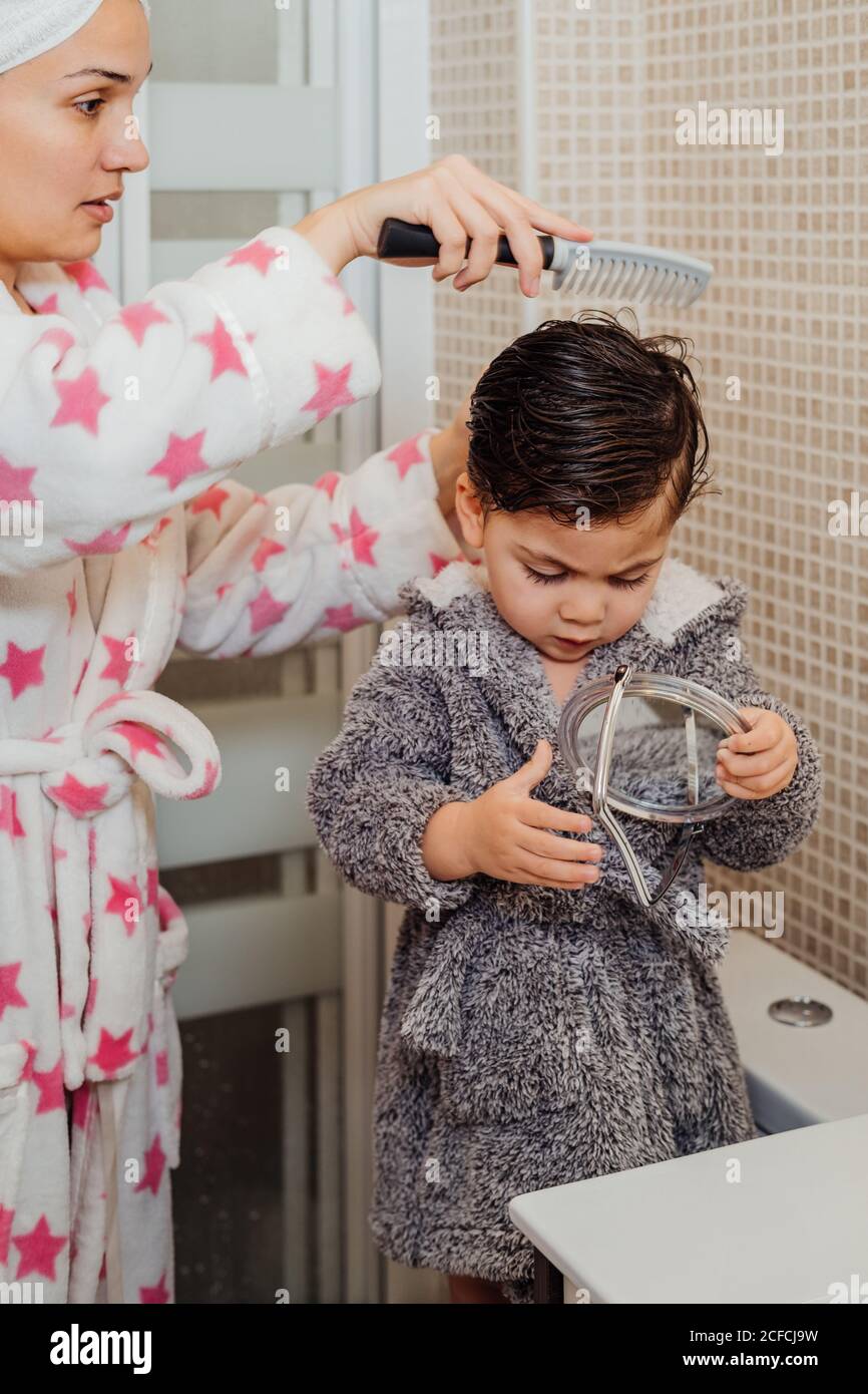 Seitenansicht der Mutter im Bademantel und Handtuch Turban Kämmen Nasse Haare von kleinen Kindern, während sie im modernen Badezimmer stehen Stockfoto