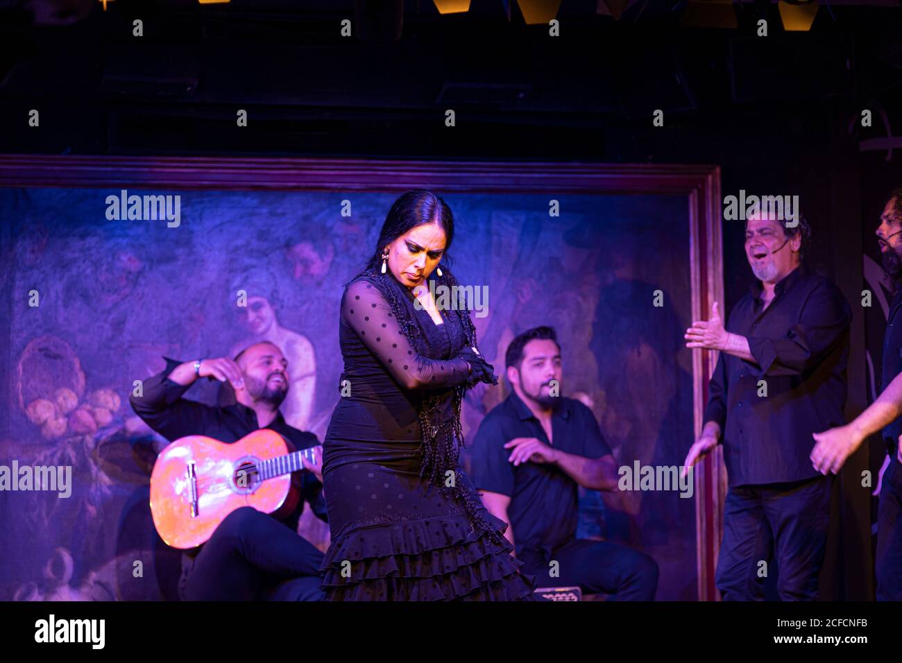 Frau in schwarzem Kostüm tanzt Flamenco in der Nähe von hispanischen männlichen Musikern Während der Performance gegen Malerei auf dunkler Bühne Stockfoto