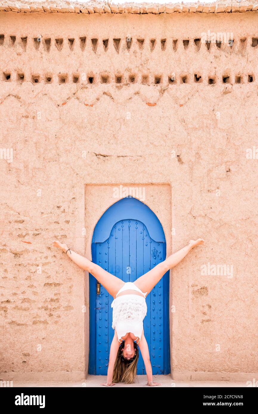 Frau in weißer Strandkleidung mit Handstand gegen orientalisch blaue Tür in Steinmauer, Marokko Stockfoto