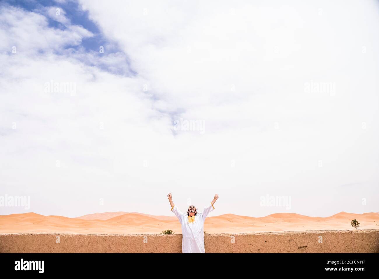 Euphorischer erwachsener Mann, der mit ausgestreckten Armen am Steinzaun auf der Terrasse gegen endlose Wüste, Marokko, den Himmel anschaut Stockfoto