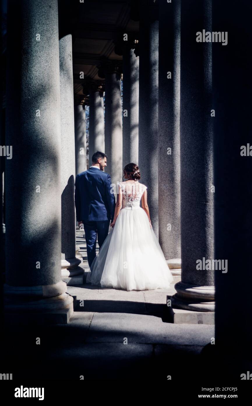 Von unten Seitenansicht des jungen eleganten Paares in der Hochzeit Kleider, die an sonnigen Tagen in der Nähe von Säulen auf der Straße spazieren Stockfoto