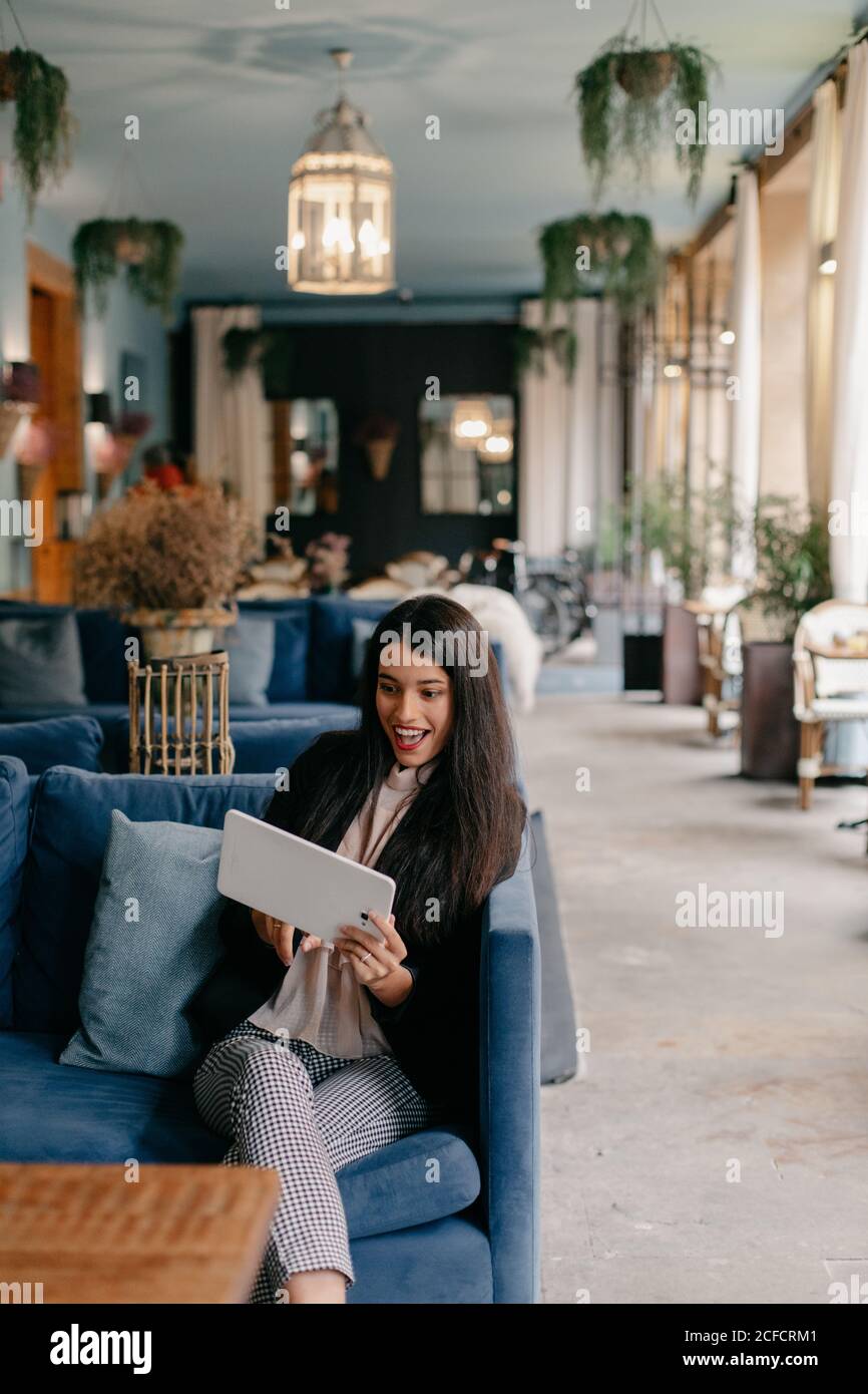 Glückliche junge Frau, die ein Tablet benutzt, während sie auf einer bequemen Couch in der Lobby sitzt Stockfoto