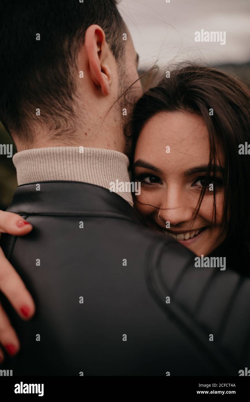 Junge Frau in der Liebe umarmt unkenntlich Freund und lächelt, während Blick auf die Kamera während des Tages Stockfoto