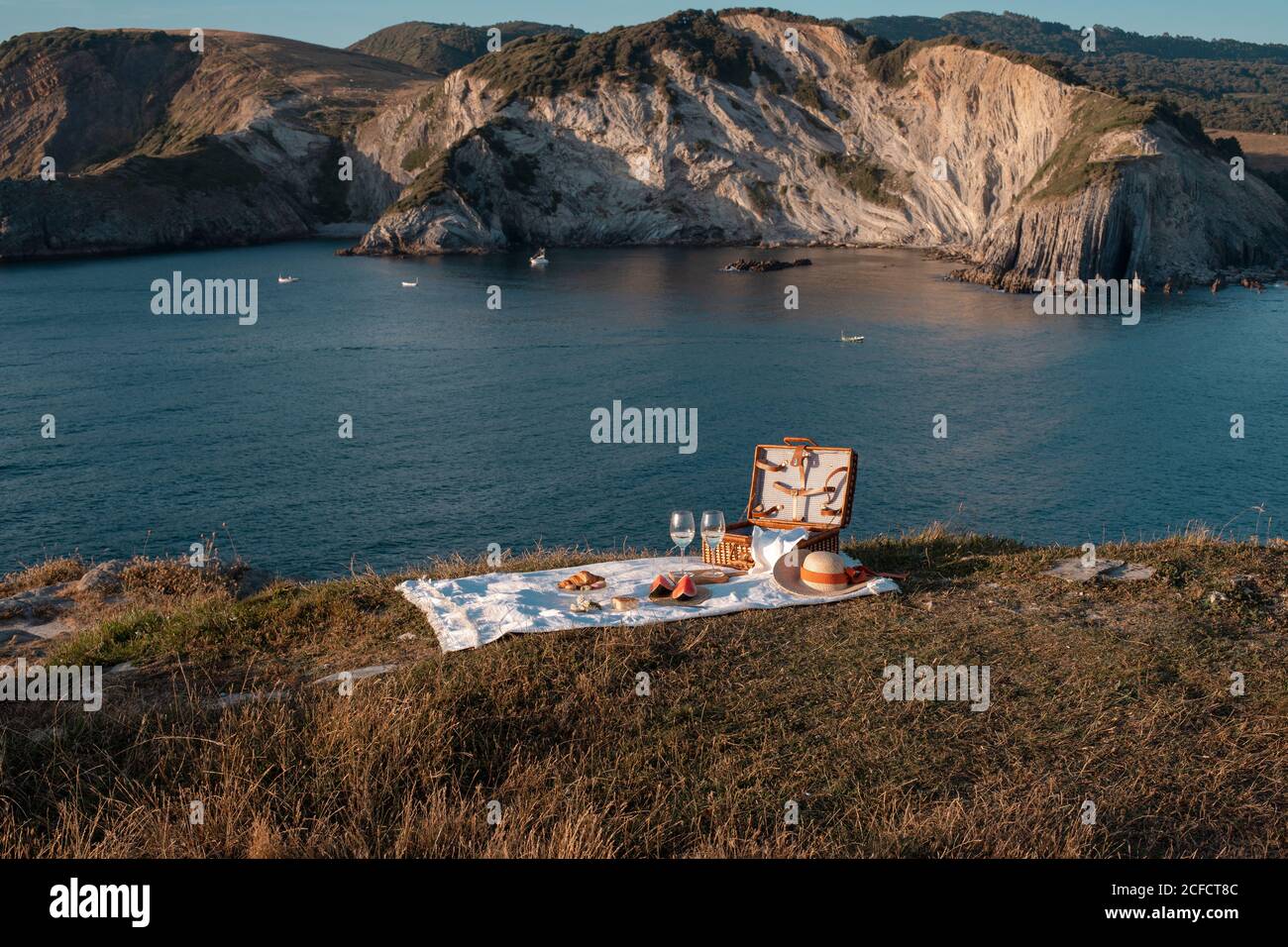 Picknickmatte mit romantischem Set mit Glas Getränk und Essen an trockener Küste mit heiterem Wasser und Hügeln Stockfoto