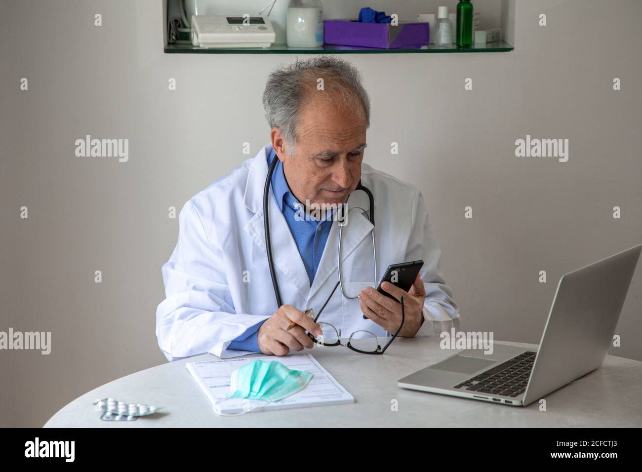 Ältere männliche Hausarzt in medizinischen Kleid sitzen am Tisch Mit Laptop in moderner Klinik und Kommunikation auf Smartphone via Videoanruf mit dem Patienten während einer Coronavirus-Pandemie Stockfoto