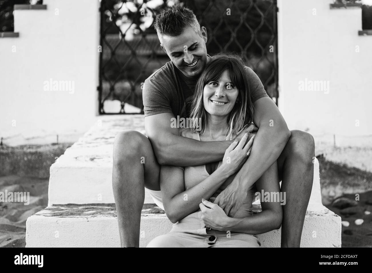 Schwarz-weiß fröhlicher erwachsener Mann und Frau, die lächeln und umarmen, während sie auf Treppen vor dem Gebäude sitzen Stockfoto