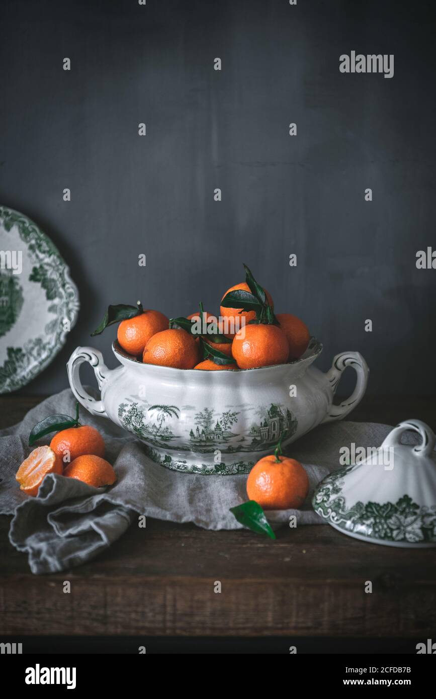 Anspruchsvolles Stillleben mit reifen orangen Mandarinen mit grünen Blättern In tiefweißer Schale auf Tisch auf grauem Hintergrund Stockfoto