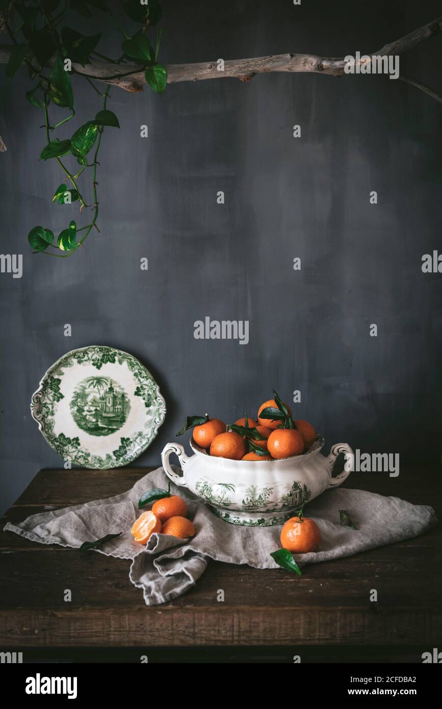 Anspruchsvolles Stillleben mit reifen orangen Mandarinen mit grünen Blättern In tiefweißer Schale auf Tisch auf grauem Hintergrund mit Baumstruktur Stockfoto
