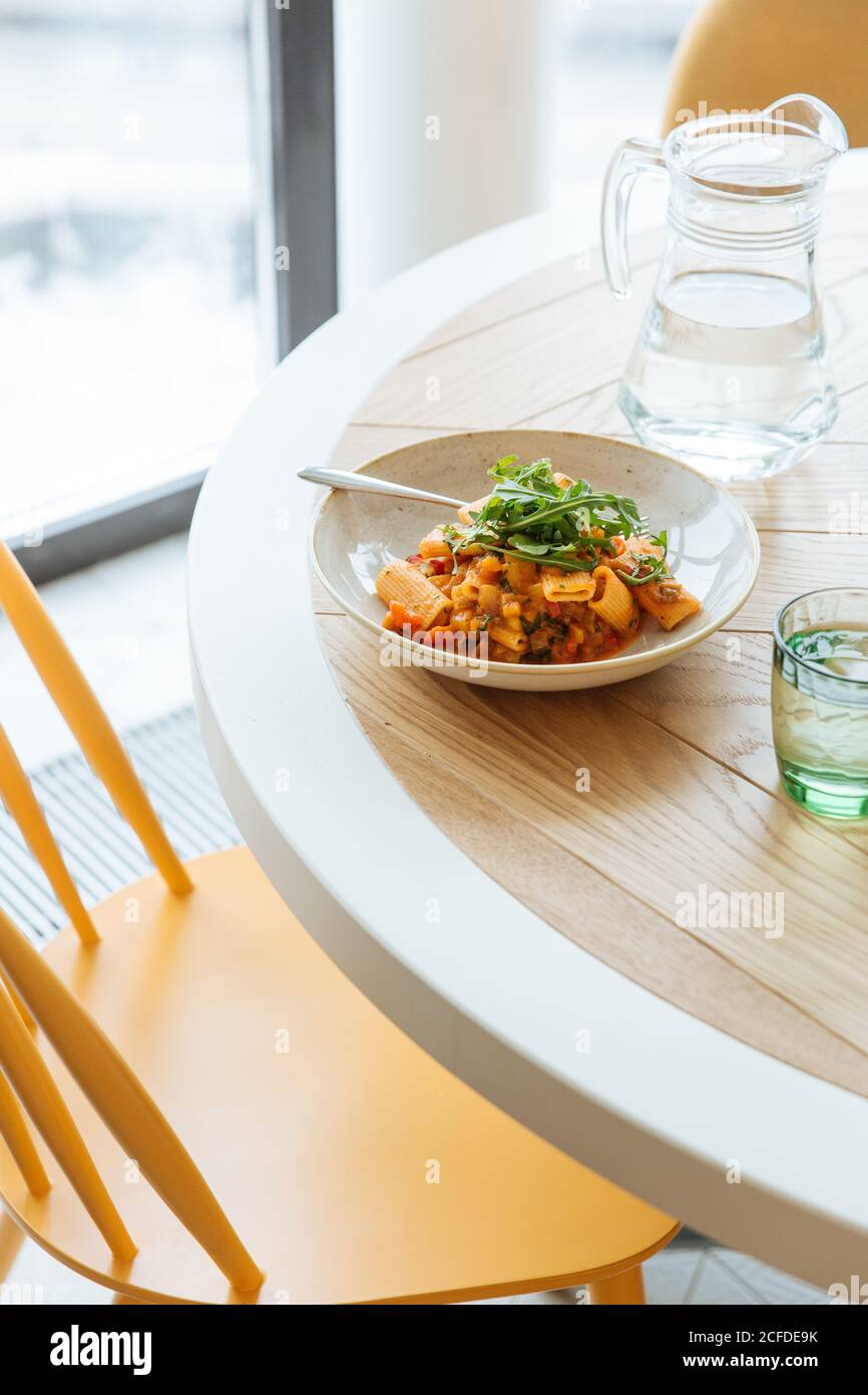 Von oben schmackhaftes buntes Ragout mit Nudel in Weiß Schüssel mit Grün auf dem Tisch mit Glas Wasser dekoriert Stockfoto