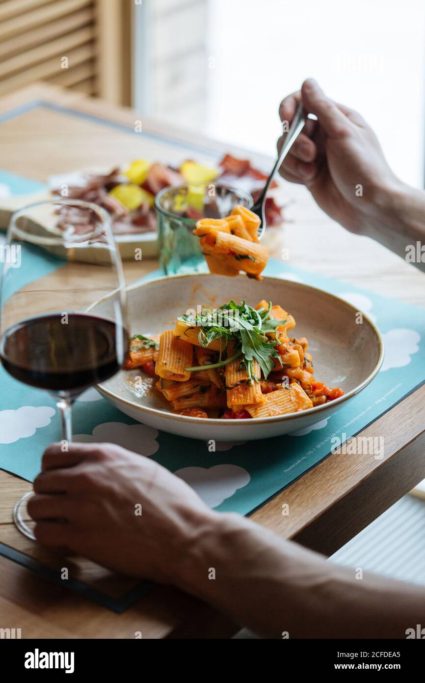 Cropped unkenntlich Person essen leckere vegane Pasta mit frischen dekoriert Rucola Blätter und Sauce und trinken Rotwein in Ein Glas an einem Restauranttisch Stockfoto