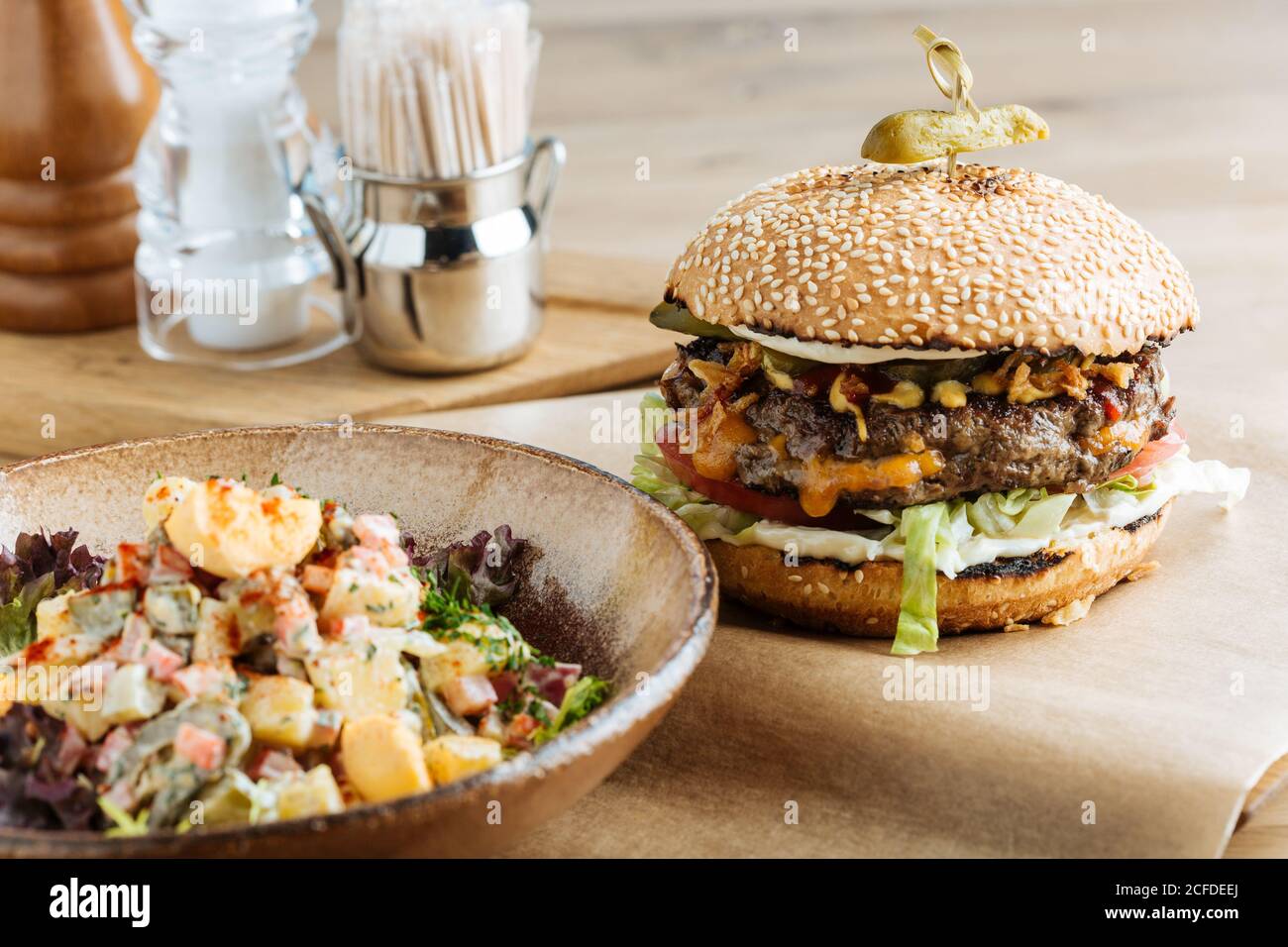 Großer Hamburger auf Papier mit Schnittkäse und Gemüse serviert Mit einer Schüssel mit buntem Salat im modernen Restaurant Stockfoto
