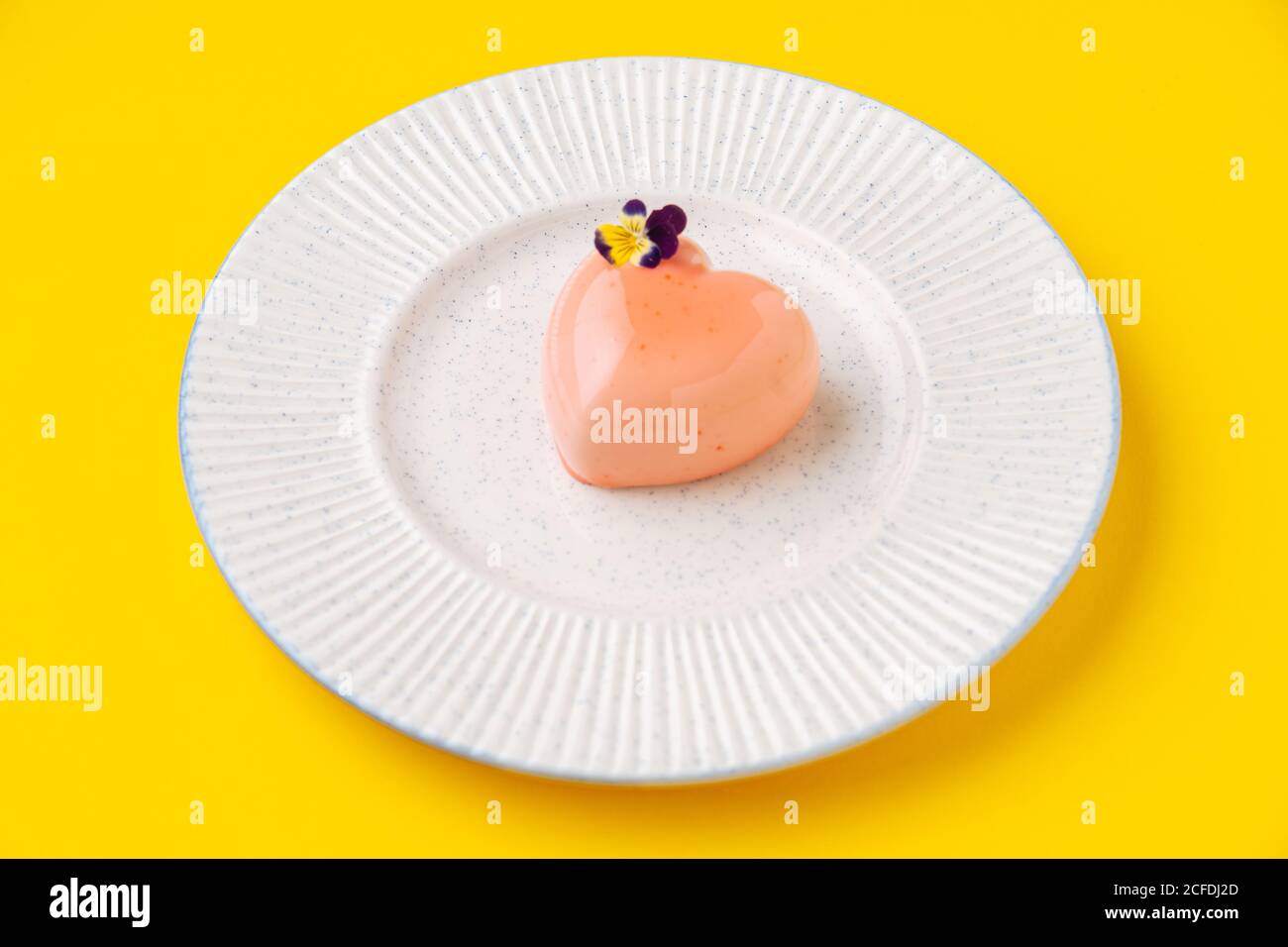 Von oben kleines herzförmiges Dessert mit rosa Vereisung und Kleine Blume auf Teller auf gelbem Hintergrund platziert Stockfoto
