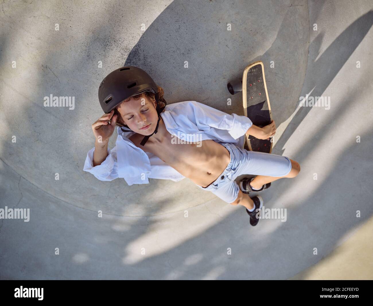 Blick von oben auf Kind im Helm liegend mit Augen geschlossen und chillen auf dem Boden im Skatepark mit Schatten Stockfoto