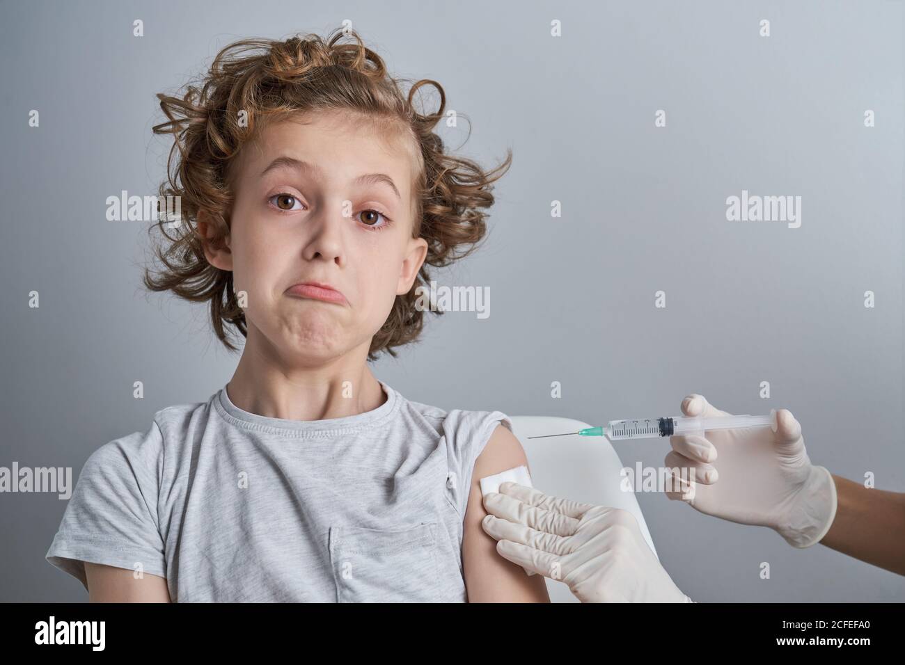 Crop Krankenschwester in weißen Latex-Handschuhe halten Schulter des Jungen Mit lockigen Haaren, während Impfstoffinjektion mit Spritze in Moderne Klinik Stockfoto