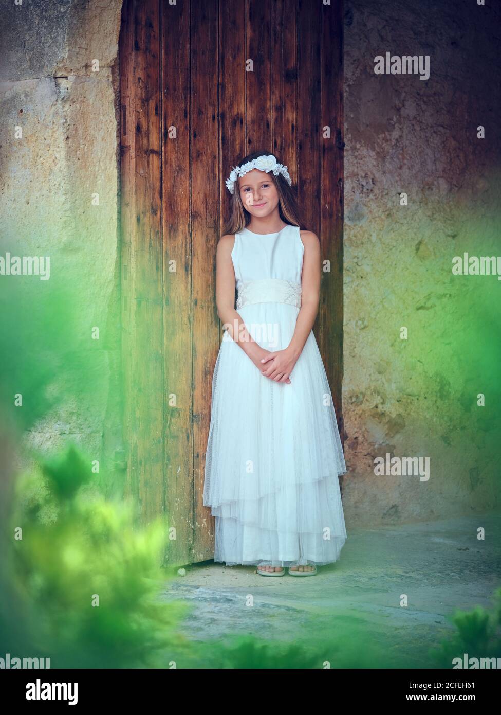 Nettes kleines Mädchen in weißen langen Kleid und Stirnband ruhen Lehnte sich an einer Holztür und Efeu schaute auf die Kamera Stockfoto