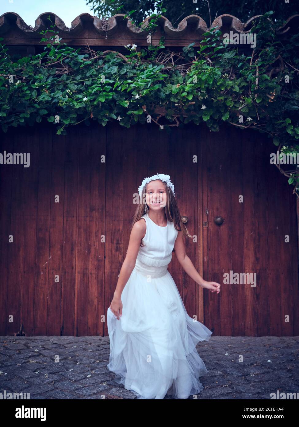Nettes kleines Mädchen in weißen langen Kleid und Stirnband springen In der Nähe einer Holztür mit Efeu, der die Kamera anschaut Stockfoto