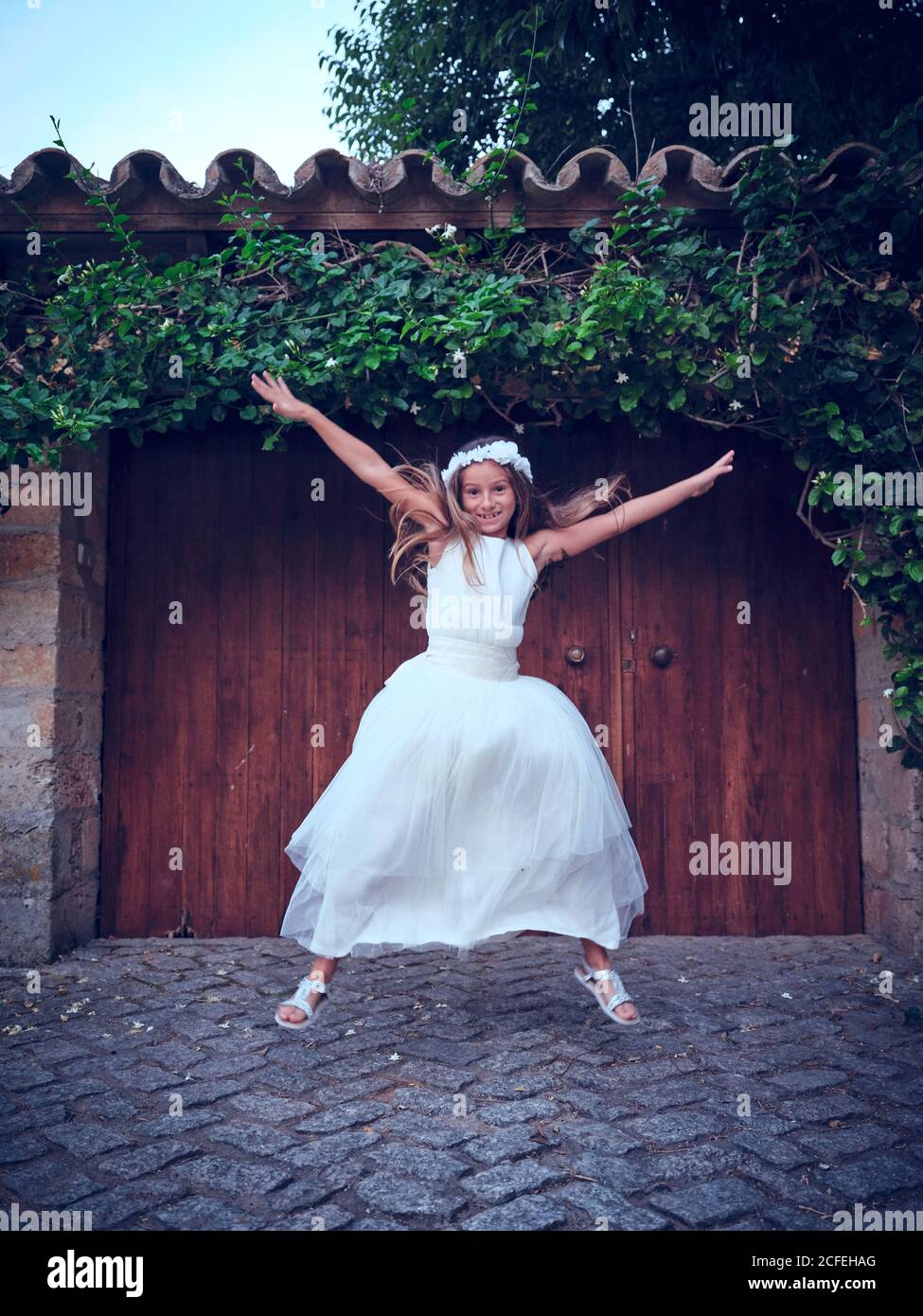 Nettes kleines Mädchen in weißen langen Kleid und Stirnband springen In der Nähe einer Holztür mit Efeu, der weg schaute Stockfoto