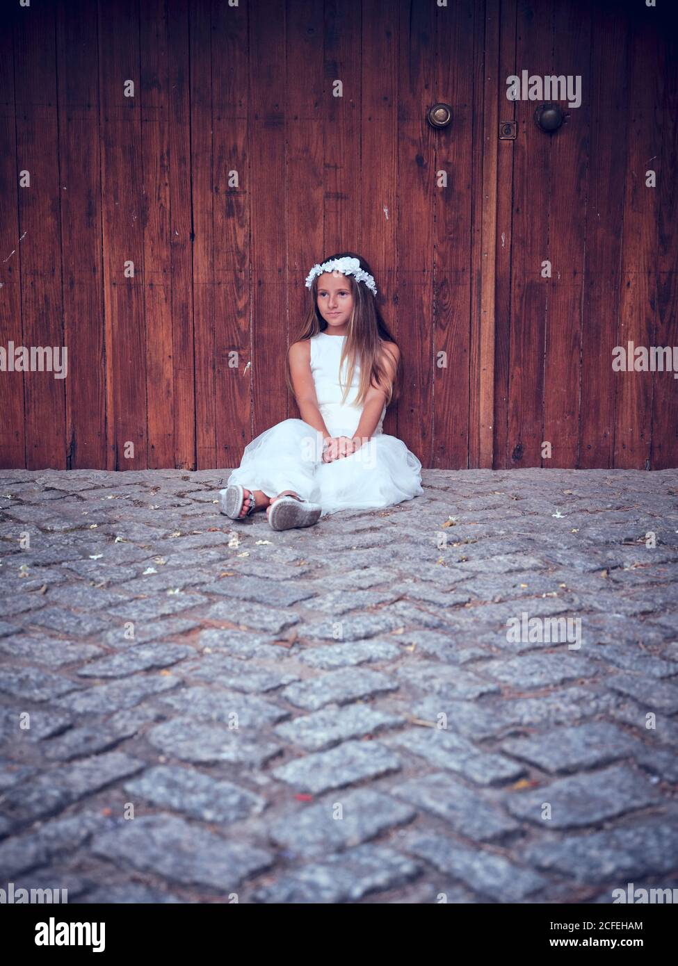 Nettes kleines Mädchen in weißen langen Kleid und Stirnband ruhen Neben der Holztür sitzend, mit Efeu, der weg schaute Stockfoto