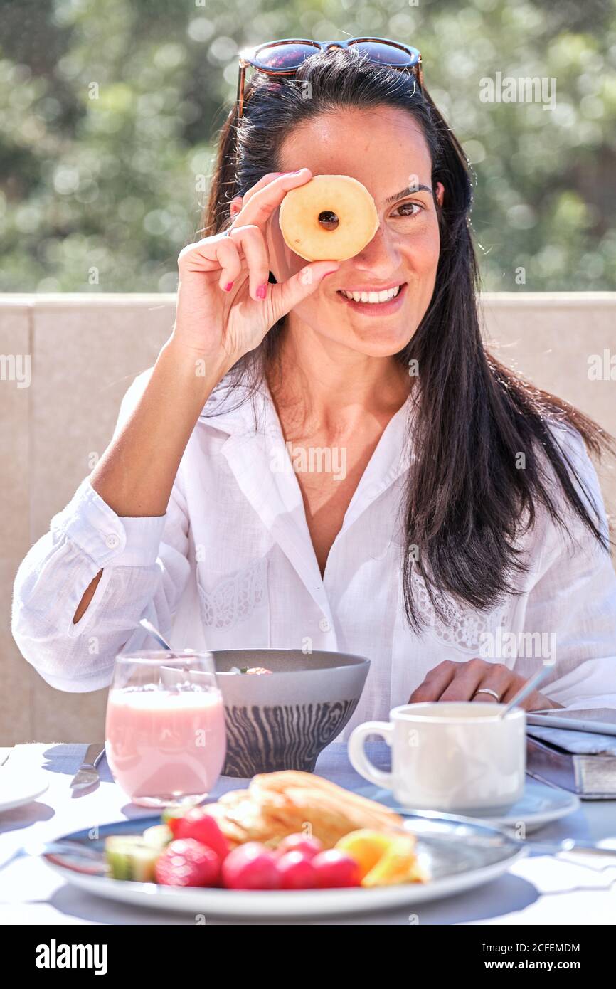 Frau, die am Tisch saß und das Frühstück auf der offenen Sonnenbeleuchtung servierte Terrasse während halten und demonstrieren Donut und lachen beim Schauen Bei der Kamera auf unscharfem Hintergrund Stockfoto