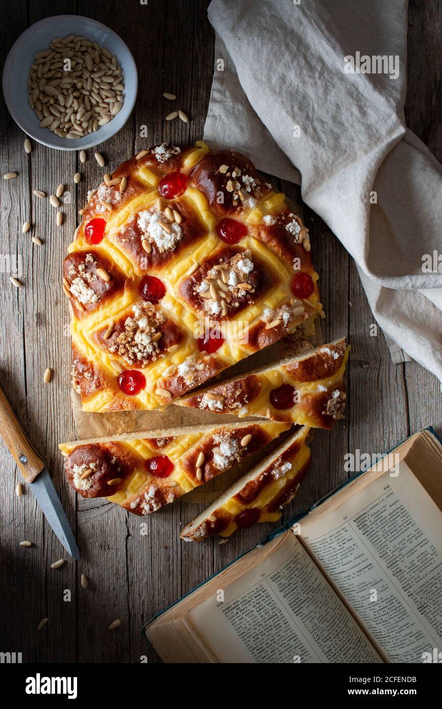 Blick von oben auf frisches Coca de San Juan Gebäck mit Samen und offenes Rezeptbuch auf Holztisch in der Nähe platziert Handtuch und Messer in rustikaler Küche Stockfoto