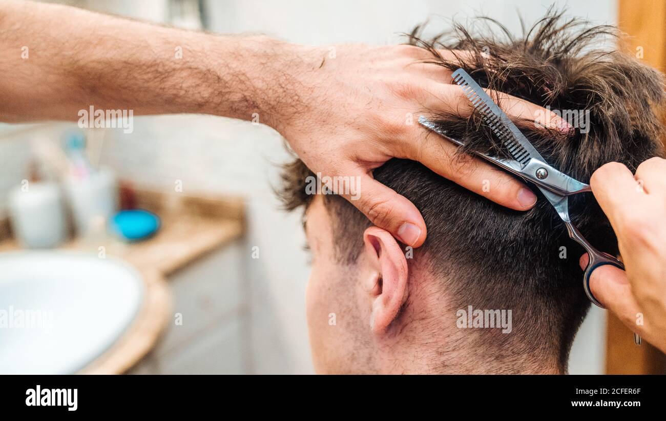 Rückansicht der unkenntlich männlichen Herstellung Haarschnitt zu Kerl mit Schere gegen verschwommenes Innenleben des hellen Badezimmers zu Hause Stockfoto