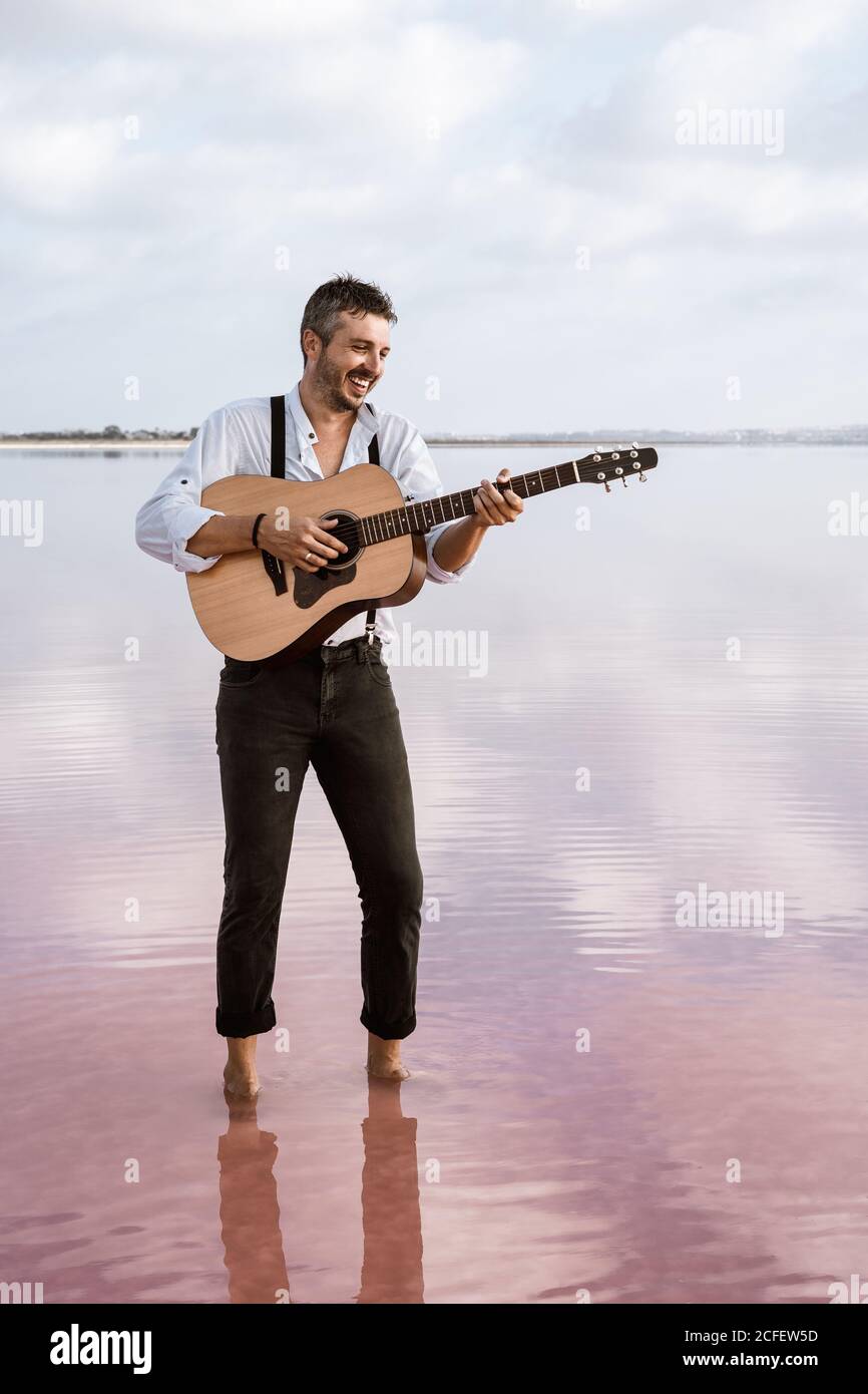 Leidenschaftlicher Mann in weißem Hemd und Hosenträger spielen Gitarre, während Barfuß im Wasser am Ufer am bewölkten Tag stehen Stockfoto