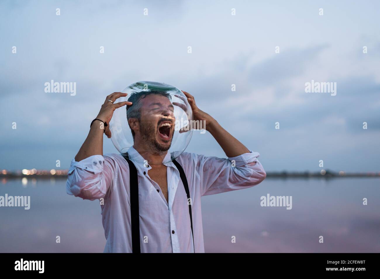 Notleidende Mann in Qual trägt nasses Hemd mit leerem Aquarium Auf dem Kopf stehend schreiend am stillen Meer in der Dämmerung Stockfoto