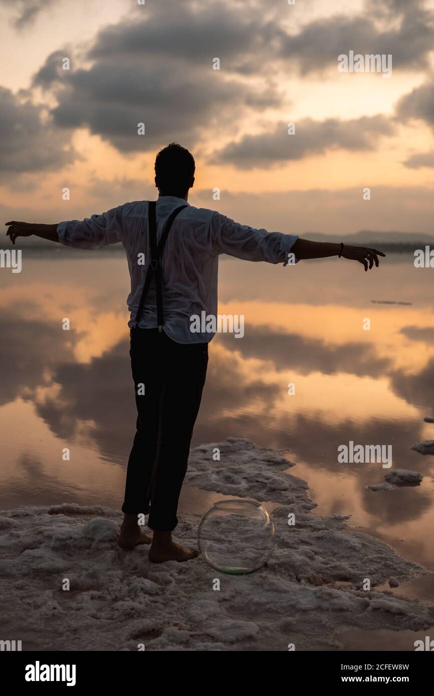 Von hinten Mann in nassen Kleidern starrt und steht mit Ausgestreckte Hände mit leerem Fischtank an der Küste bei Wolken Sonnenuntergang Stockfoto