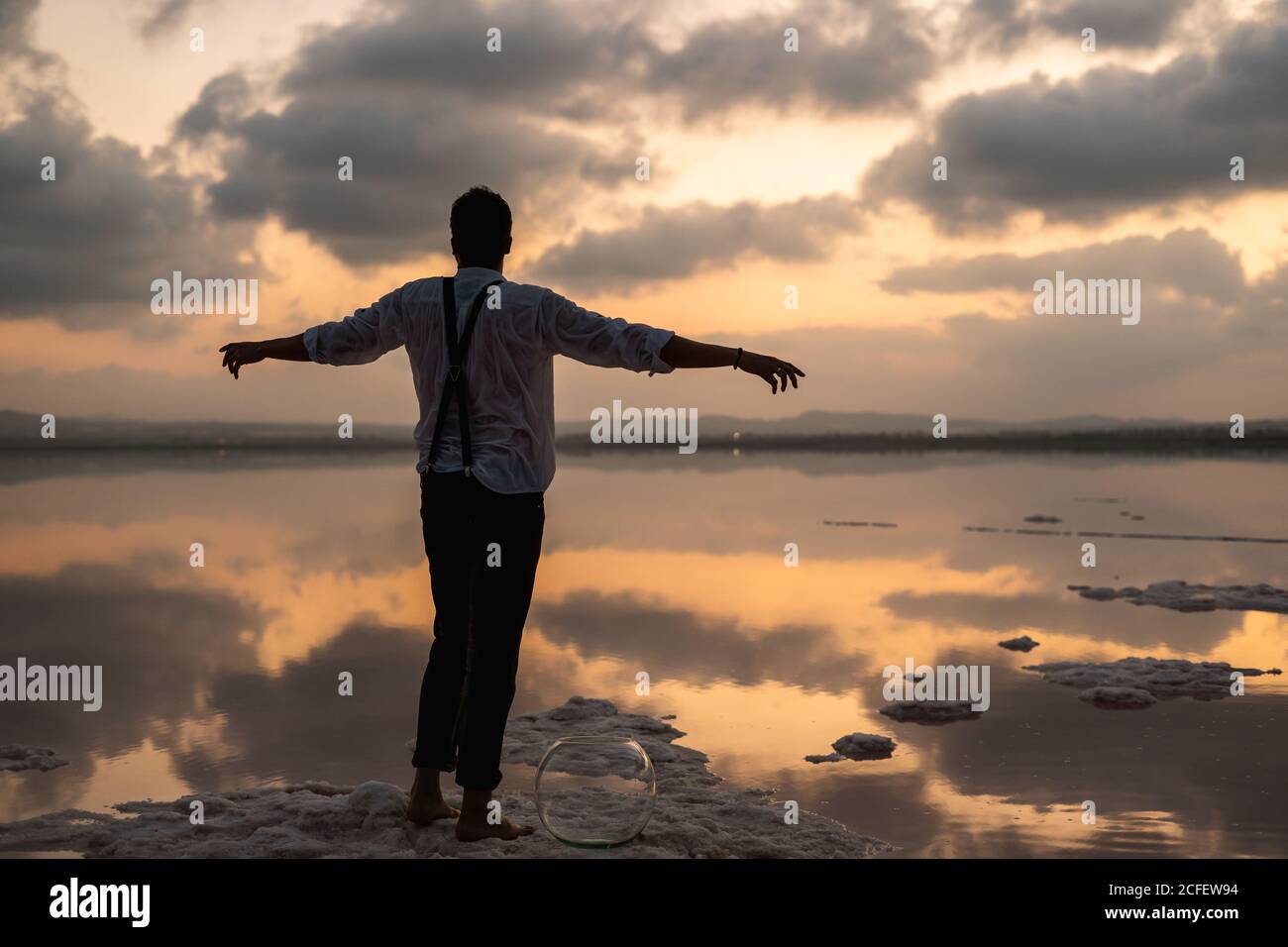 Von hinten Mann in nassen Kleidern starrt und steht mit Ausgestreckte Hände mit leerem Fischtank an der Küste bei Wolken Sonnenuntergang Stockfoto