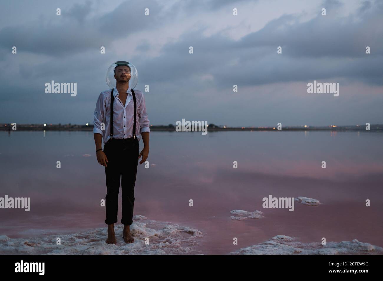 Nachdenklicher Mann mit geschlossenen Augen im nassen Hemd mit leerem Aquarium auf dem Kopf stehend und kontemplierend durch stilles Meer in Dämmerung Stockfoto