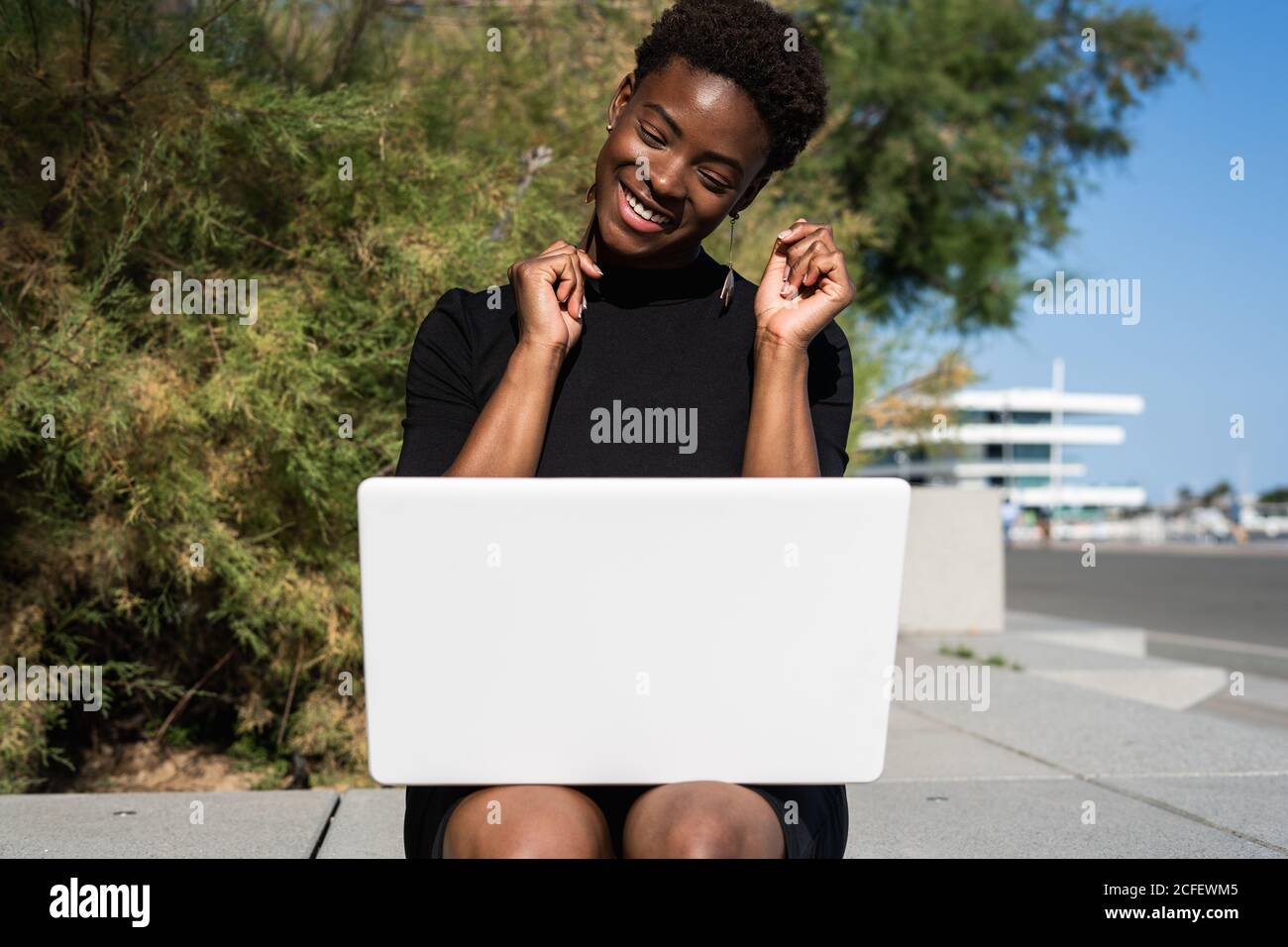 Fröhlich aufgeregt Frau in elegantem schwarzen Kleid mit Laptop, während auf dem Bürgersteig auf der Straße entspannen Stockfoto