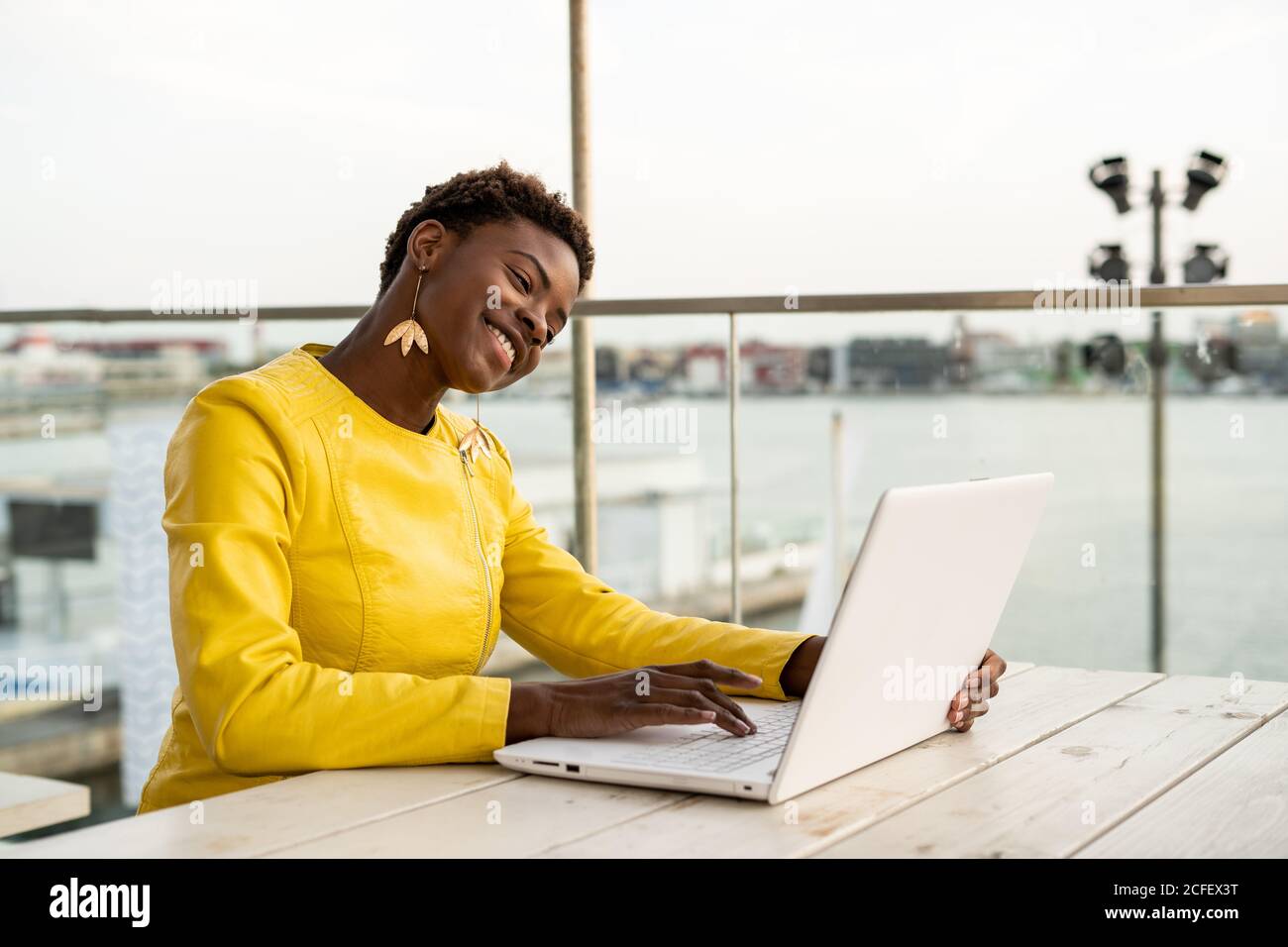 Schwarze afroamerikanische Frau in gelber Jacke mit Laptop-Webcam am Holzschreibtisch in der Stadt auf verschwommenem Hintergrund Stockfoto