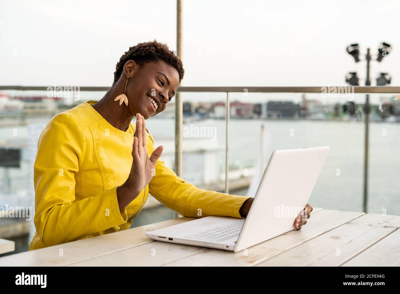 Schwarze afroamerikanische Frau in gelber Jacke mit Laptop-Webcam am Holzschreibtisch in der Stadt auf verschwommenem Hintergrund Stockfoto