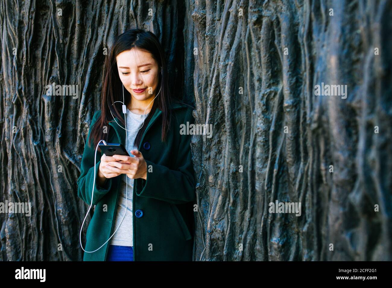 Schöne asiatische Frau in stilvollem Outfit, Musik zu hören und mit Smartphone, während sie sich an rauer Wand mit Relief von Baumrouten lehnt Stockfoto