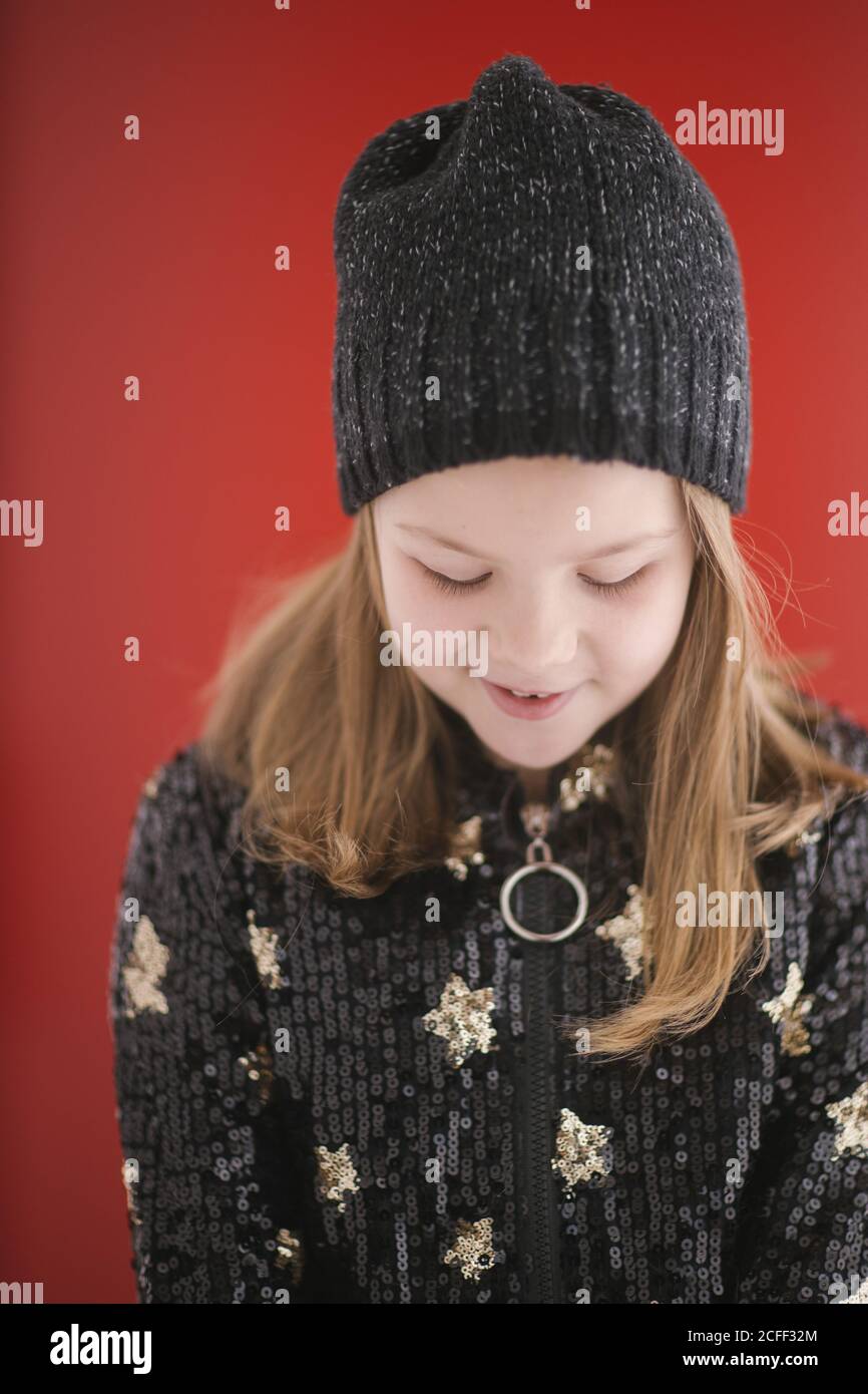 Fröhliches Kind mit toothy Lächeln in warmen schwarzen Jersey und Gestrickte Mütze mit Blick nach unten auf rotem Hintergrund Stockfoto