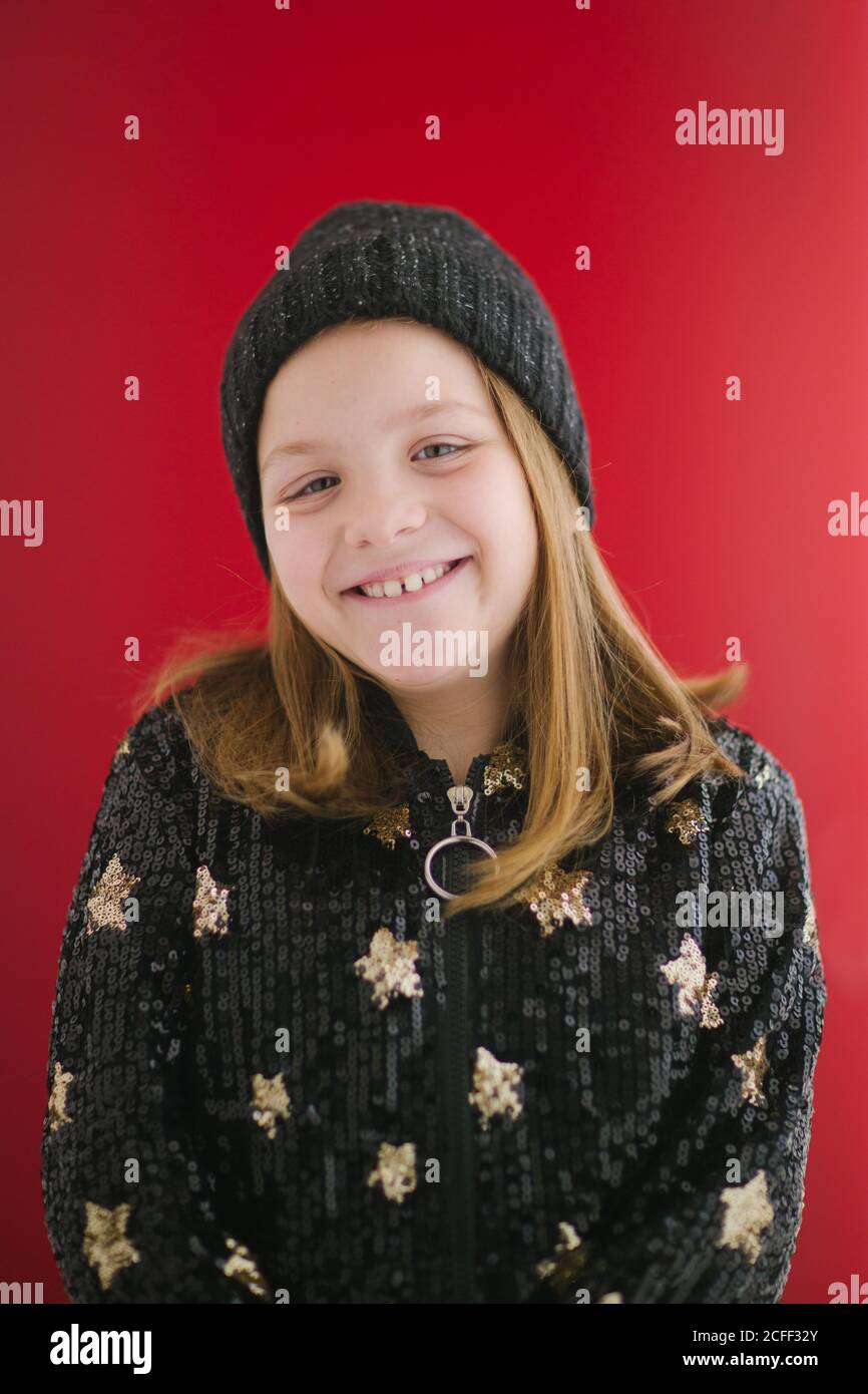 Fröhliches Kind mit toothy Lächeln in warmen schwarzen Jersey und Strickmütze, die auf rotem Hintergrund auf die Kamera schaut Stockfoto