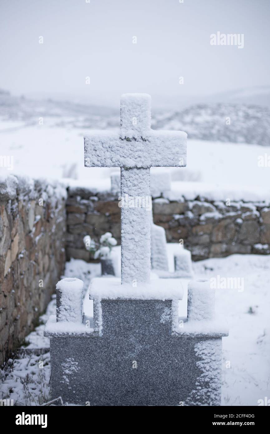 Schnee bedeckt Alter graues Kreuz Grabstein in der Nähe Zaun mit wolkiger grauer Himmel und weiße Felder auf dem Hintergrund Stockfoto