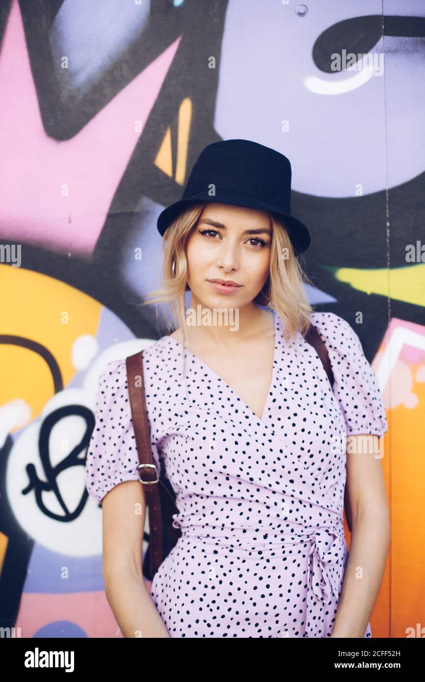 Porträt einer selbstbewussten jungen Frau in Kleid und schwarzem Hut, die auf die Kamera an der Graffiti-Wand blickt Stockfoto