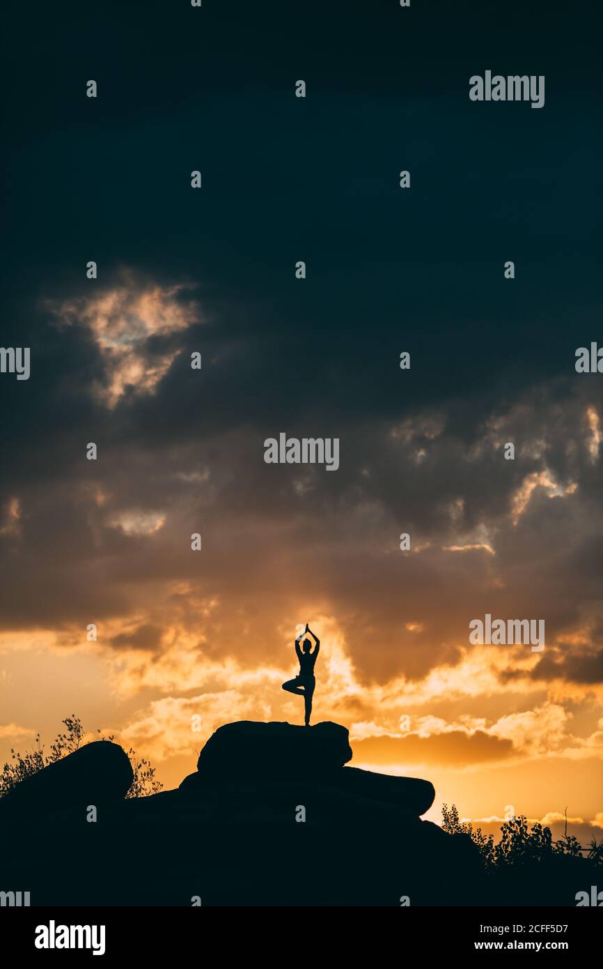 Silhouette einer Frau, die Yoga macht, posiert im Freien bei einem atemberaubenden Sonnenuntergang, während sie auf dem Felsen steht Stockfoto