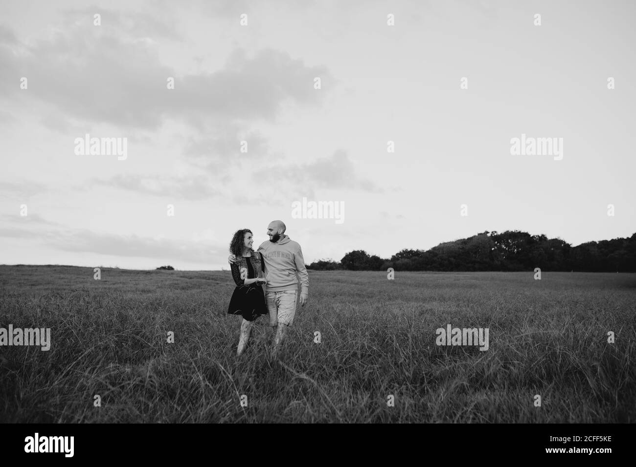 Fröhliches romantisches junges Paar, das Hände hält und auf Grün läuft Feld am Meer bei Sonnenuntergang mit bewölktem Himmel Stockfoto