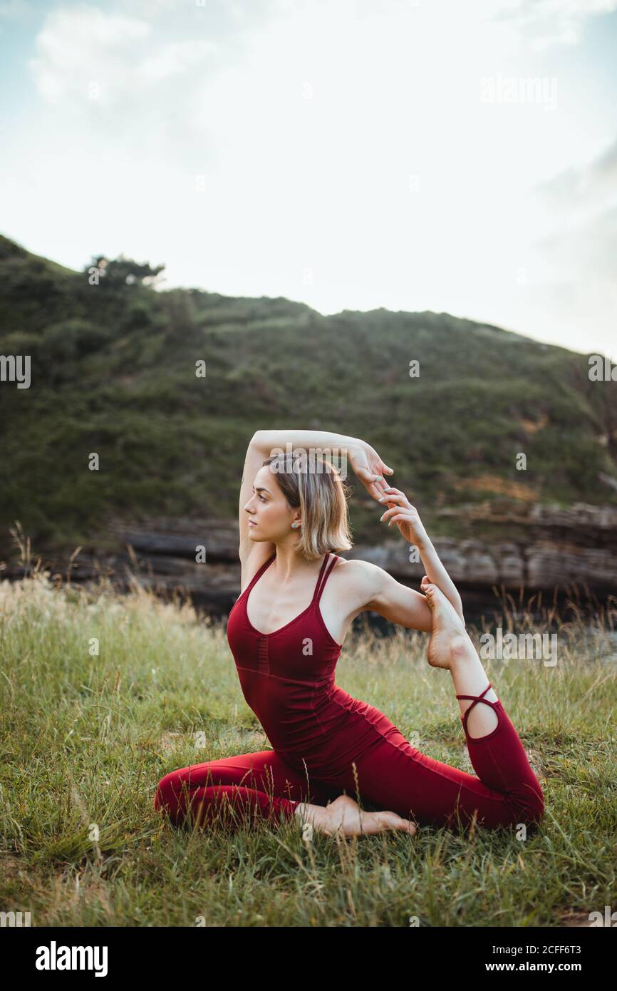 Junge flexible Frau in roter Sportkleidung sitzt in schwierigen Yoga einbeinigen Königstaube Pose auf leeren grünen Hügel Stockfoto