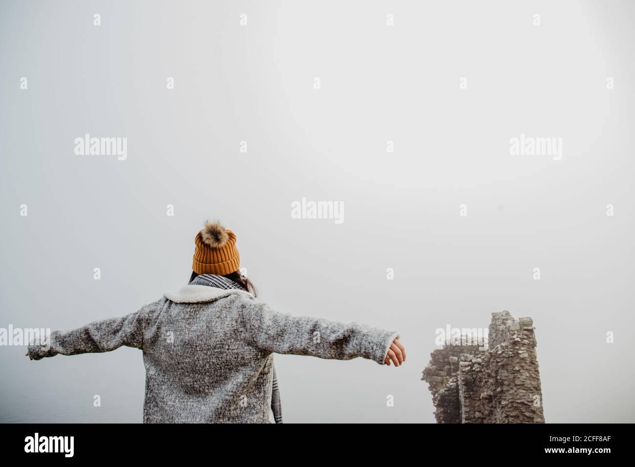 Rückansicht einer anonymen Frau in warmer Kleidung mit offenen Armen, die am bewölkten Tag im nebligen Park spazieren geht Stockfoto