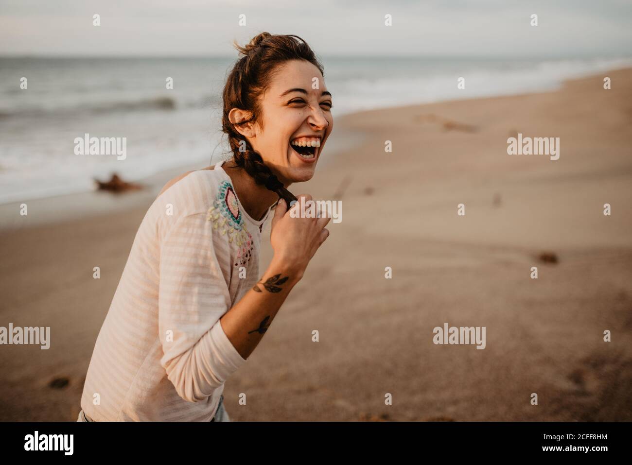 Junge Dame im lässigen Outfit berühren Zopf und lachend aus Laut beim Stehen an der sandigen Küste Stockfoto